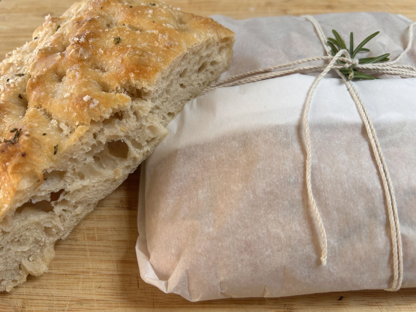 rosemary focaccia cut in half resting on a loaf wrapped in parchment paper, tied with twine with a sprig of rosemary